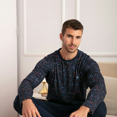 Man sitting on a bed wearing a dark blue patterned long-sleeve shirt and dark pants.